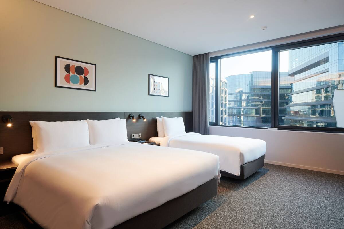 Ein Hotelzimmer mit zwei Betten, einem großen Fenster mit Blick auf die Stadt und Bildern an der Wand.
