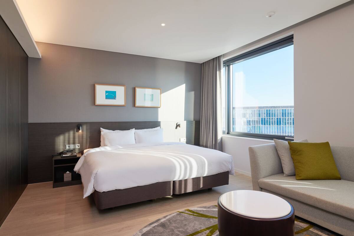Ein modernes Hotelzimmer mit einem großen Bett, einer Couch, einem kleinen Tisch und einem Fenster mit Blick auf ein Gebäude.
