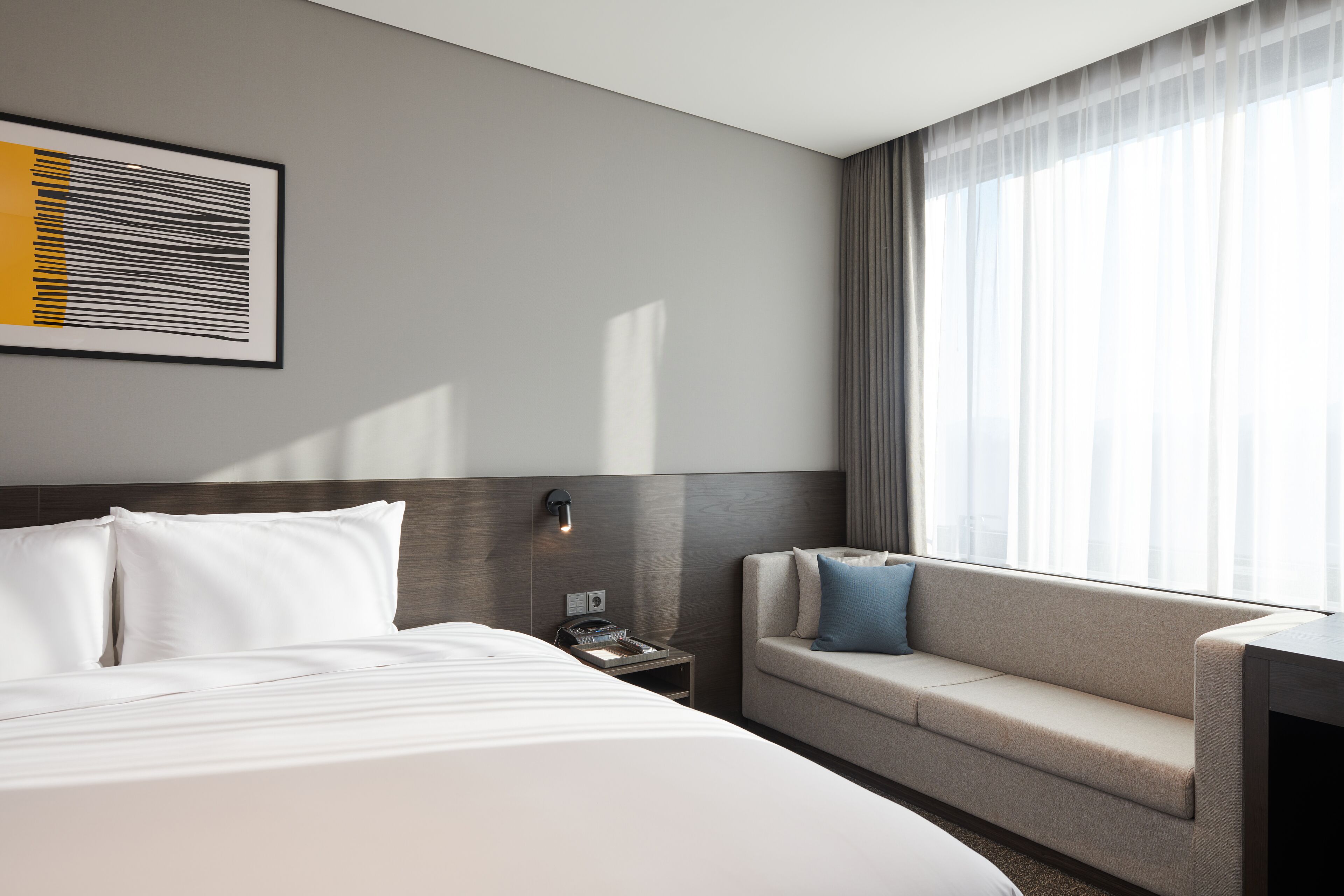 A modern hotel room with a large bed, a sofa, a nightstand, and a wall-mounted phone.