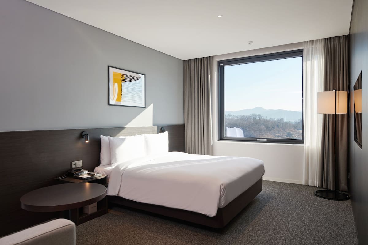Ein modernes Hotelzimmer mit einem großen Bett, einem Nachttisch, einem kleinen runden Tisch, einer Wandlampe und einem großen Fenster mit Bergblick.