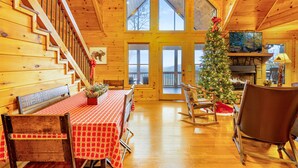 Ein Holzblockhaus-Interieur mit einem Esstisch, Stühlen, einem Schaukelstuhl, einem Weihnachtsbaum und einem Kamin.