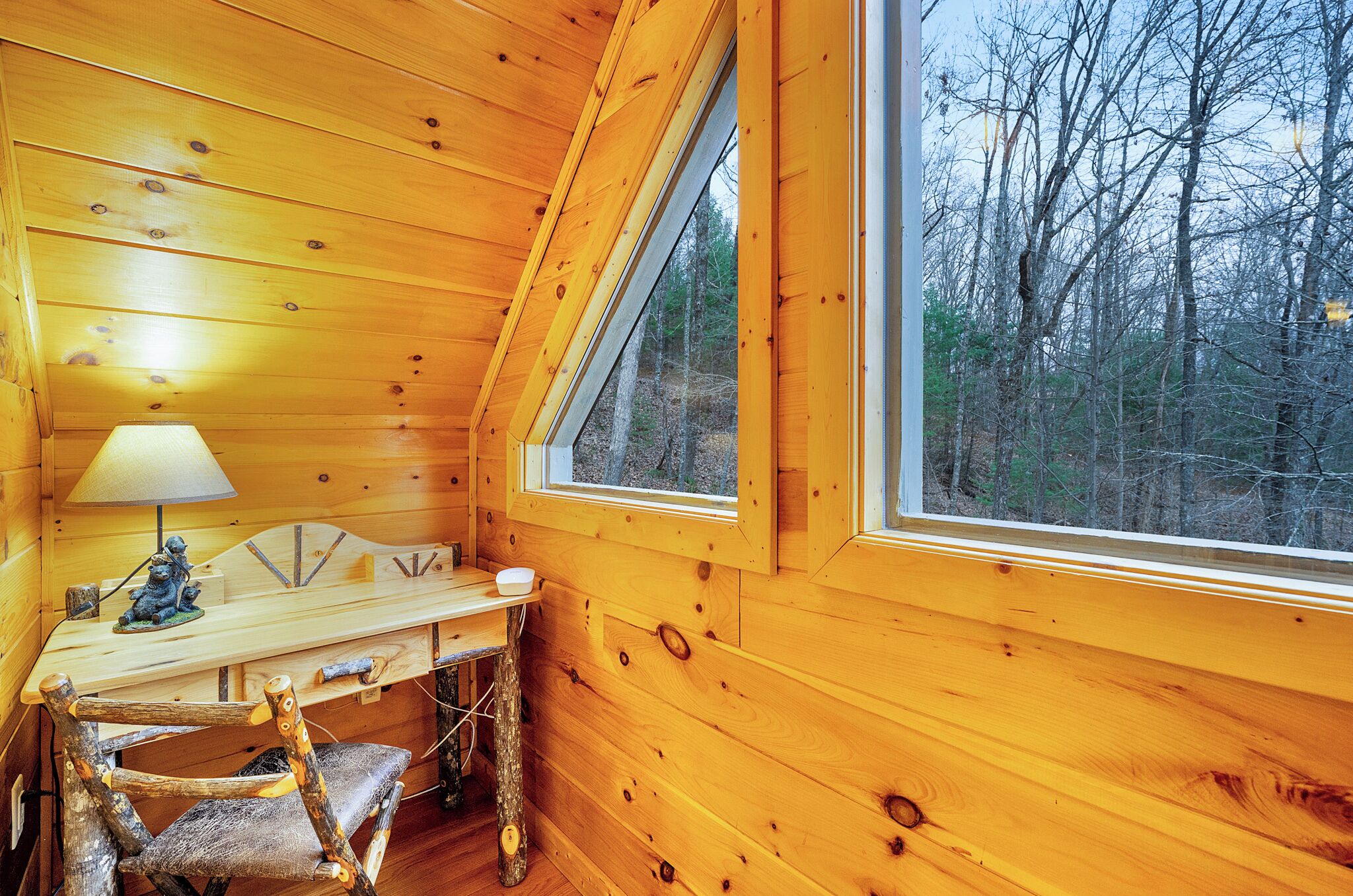 A wooden cabin interior with a desk, chair, lamp, and a large window overlooking a forest.
