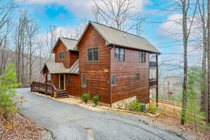 A two-story wooden cabin with a porch, a stone foundation, and a view of the surrounding forest.