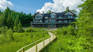A large, multi-story building with a prominent balcony area, surrounded by lush greenery and a wooden walkway leading to the entrance. - Camp Balsam | Lake Placid Lakefront Estate with Indoor Pool & Boathouse (Lake Placid)