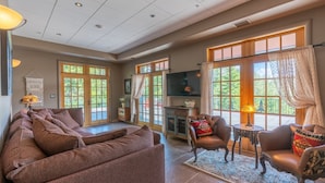 A spacious living room with a large sectional sofa, a leather armchair, and a small coffee table. There are large windows with curtains, a flat-screen TV, and a macrame wall hanging. - Camp Balsam | Lake Placid Lakefront Estate with Indoor Pool & Boathouse (Lake Placid)