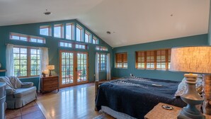 A spacious bedroom with a large bed, a wooden dresser, a chair, and a desk with a lamp. There are multiple windows providing natural light and a view of the outdoors. - Camp Balsam | Lake Placid Lakefront Estate with Indoor Pool & Boathouse (Lake Placid)