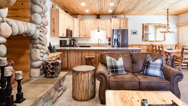 Un acogedor interior de cabaña con una pared de piedra, techo de madera, un sofá de cuero marrón y una zona de cocina con armarios de madera y un refrigerador.