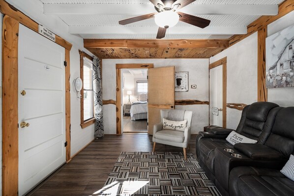 A hallway with a wooden ceiling, a black leather sofa, a patterned rug, and a chair with a cushion.