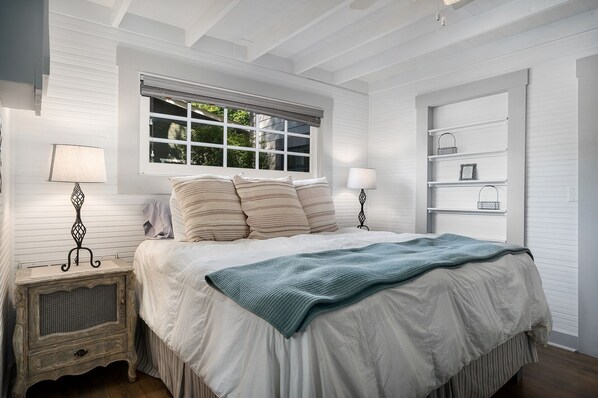 A bedroom with a large bed, two bedside tables with lamps, a window with blinds, and a built-in shelving unit. - Bowie Cottage | Hot Tub | 2 Fireplaces (Fredericksburg)