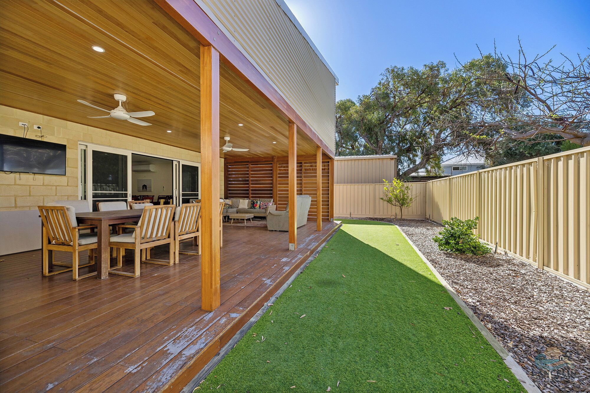 A covered patio with wooden flooring, a dining area with chairs and a table, and a small lawn with mulch.
