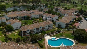 Un quartier résidentiel composé de plusieurs bâtiments blancs aux toits de tuiles, avec une piscine centrale et, au loin, un terrain de golf.