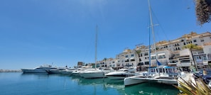 Uma marina com vários iates atracados, um hotel chamado 'BENABOLA' com varandas, e um céu azul límpido.