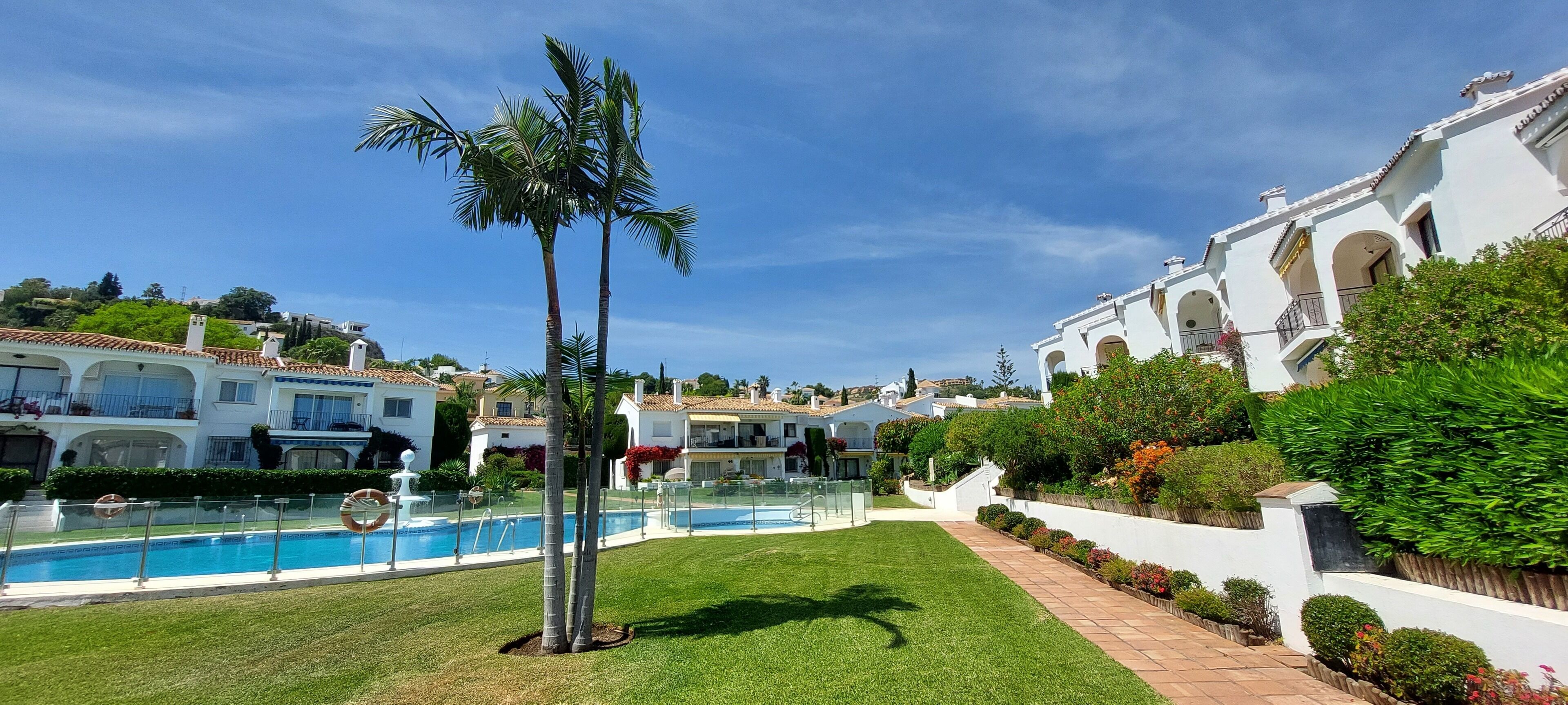 A pool surrounded by well-maintained grass and palm trees, with white buildings featuring arched windows and balconies in the background.