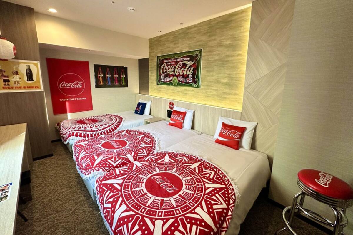 Une chambre d’hôtel avec trois lits, chacun recouvert d’une couvre-lit à motifs rouges et blancs, une affiche Coca-Cola accrochée au mur et un tabouret de bar avec un siège rouge.