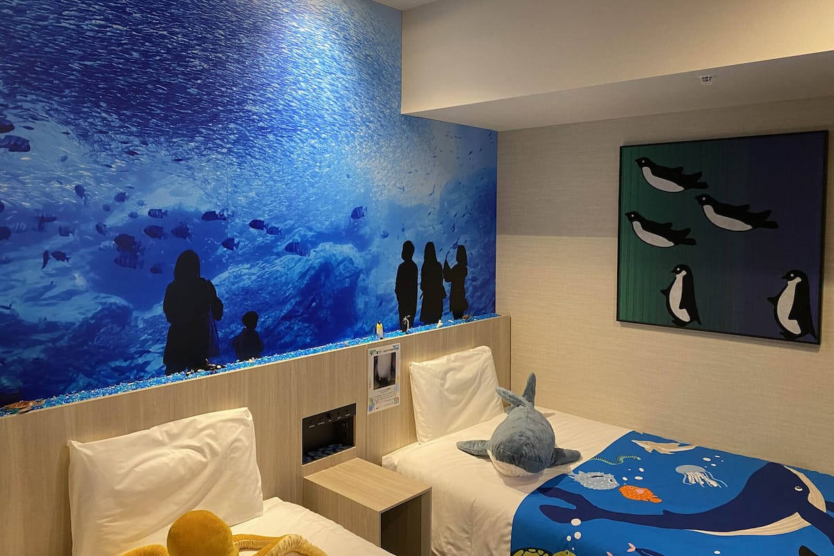 Une chambre d’hôtel avec un grand mur transformé en aquarium, un lit recouvert d’une courtepointe aux motifs marins bleus, un gros jouet en forme de requin et un tableau représentant des pingouins.