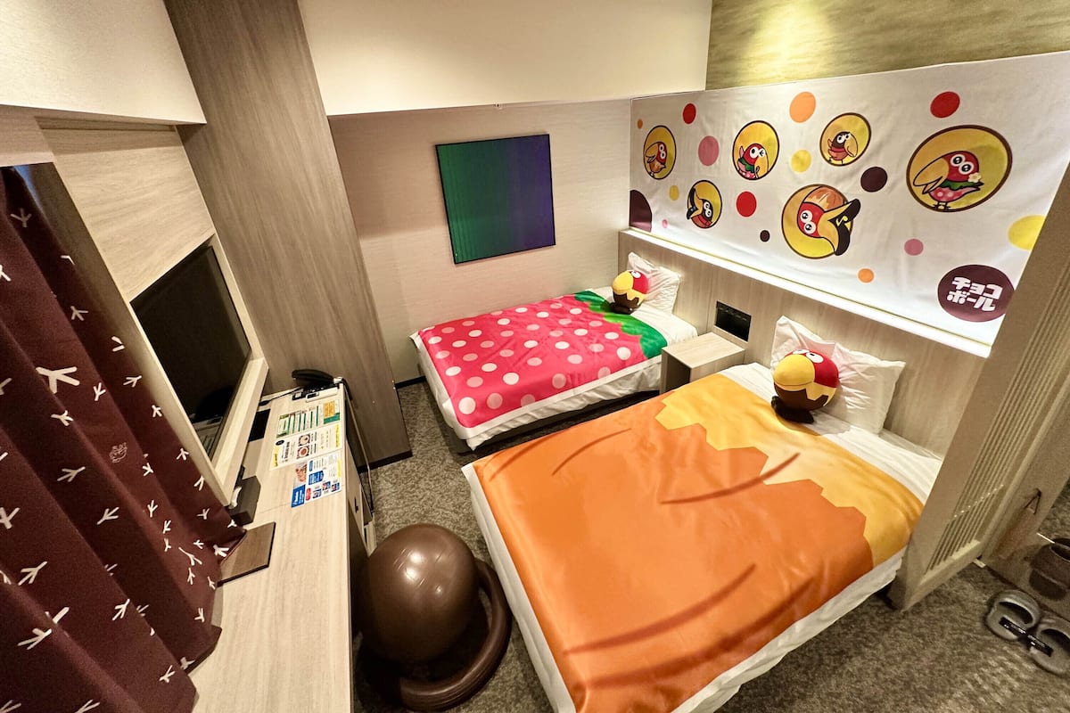 Une chambre d’hôtel avec deux lits, un bureau, une chaise, un petit ballon d’exercice et un mur décoré de personnages de dessins animés.