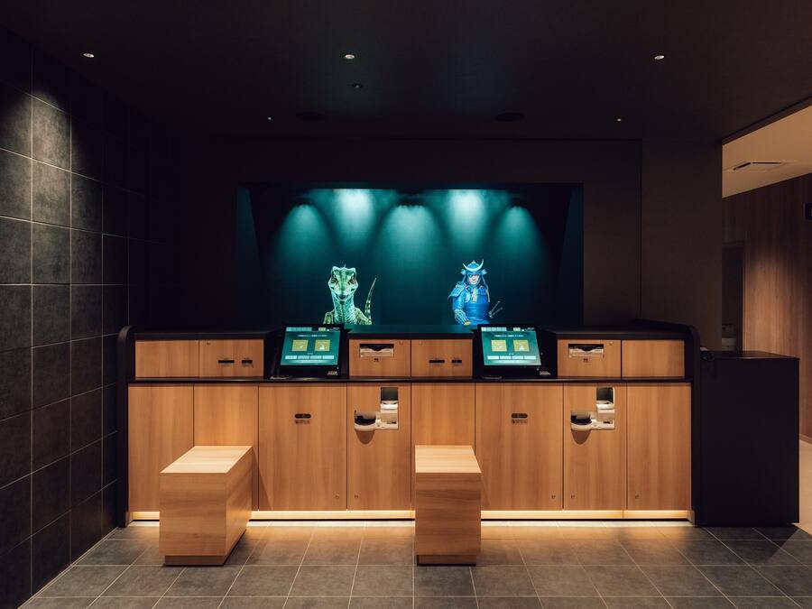 A reception area with a wooden counter, two digital kiosks, and a large screen displaying a character.