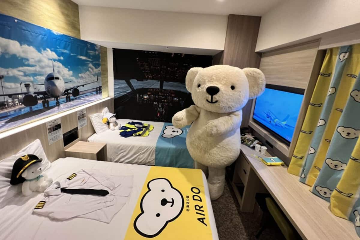 Une pièce décorée avec une grande peluche en forme d’ours, un lit avec un oreiller à motifs d’ours, un chapeau de capitaine et un écran de simulateur de vol.