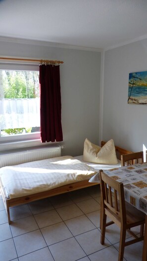 Ein kleiner, ordentlicher Raum mit einem Einzelbett, einem Stuhl, einer Heizung und einem Fenster mit Vorhängen.