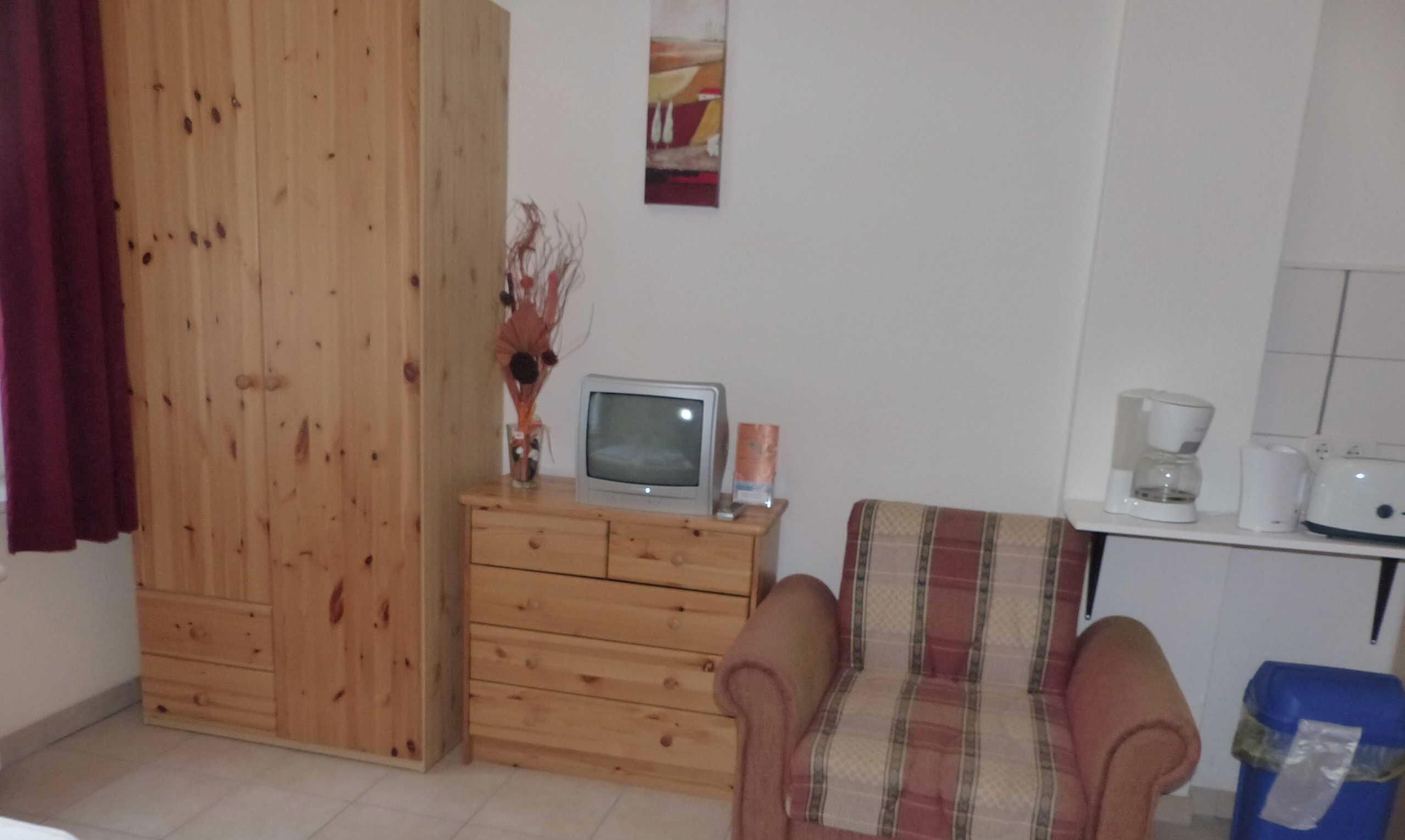 A small, simple room with a wooden wardrobe, a television on a wooden dresser, a plaid chair, and a coffee maker on a counter.