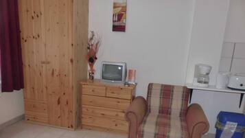 A small, simple room with a wooden wardrobe, a television on a wooden dresser, a plaid chair, and a coffee maker on a counter.