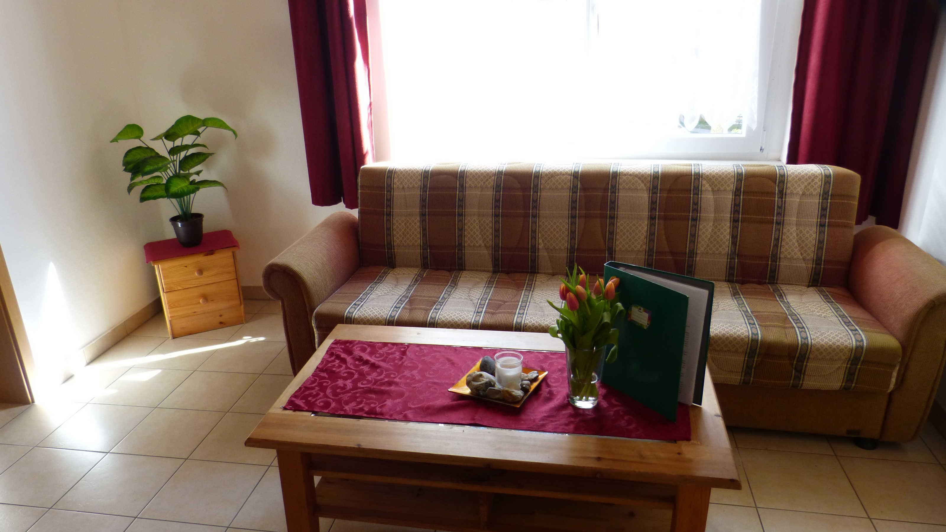 Ein Wohnzimmer mit einer Couch, einem Holztisch, einem kleinen Beistelltisch mit Pflanze und einem Fenster mit Vorhängen.