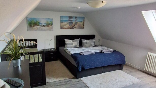A bedroom with a sloped ceiling, a bed with a blue blanket, a desk with a plant, and artwork on the walls.