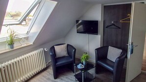 A modern room with a sloped ceiling, a radiator, a TV mounted on the wall, two black chairs, a small table, and a door with a coat rack.