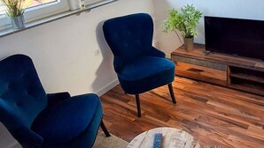 A living room with two blue armchairs, a round coffee table with a remote control, a wooden TV stand, and a potted plant.
