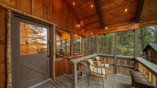Eine Holzterrasse mit einer Bank und Stühlen, einer Tür und großen Fenstern mit Blick auf einen Wald.
