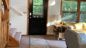 A log cabin interior with a black door, a staircase, a sofa, and a window overlooking greenery. - Søsterhus, Three Lakes/Eagle River Lake House (Eagle River)