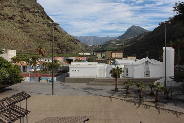 A church with a white facade, a bell tower, and a surrounding area with palm trees and a mountainous backdrop.
