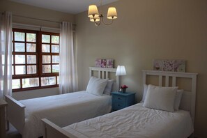 A bedroom with two beds, a window with curtains, a nightstand with a lamp, and wall art.