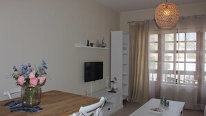 Ein modernes Wohnzimmer mit einem Holzesstisch, weißen Stühlen, einem an der Wand befestigten Fernseher, einem weißen Schrank und einer Pendelleuchte.