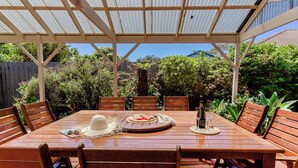 A covered outdoor dining area with a wooden table and chairs, surrounded by lush greenery and a wooden fence. - Beach Pad - 4pm check out Sundays* (Safety Beach)