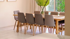 A dining area with a wooden table and chairs, a large plant, and a window with curtains. - Beach Pad - 4pm check out Sundays* (Safety Beach)