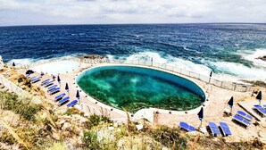 Een zwembad aan de kust met een gebogen rand, omringd door ligbedden en parasols, met uitzicht op de oceaan.