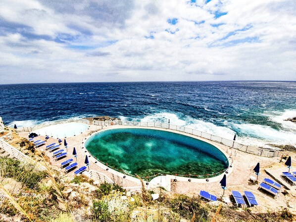 Ein Küsten-Schwimmbecken mit gewölbter Kante, umgeben von Liegestühlen und Sonnenschirmen, mit Blick auf den Ozean.
