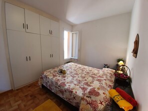 A bedroom with a bed, a window, a wardrobe, and a nightstand with a lamp.