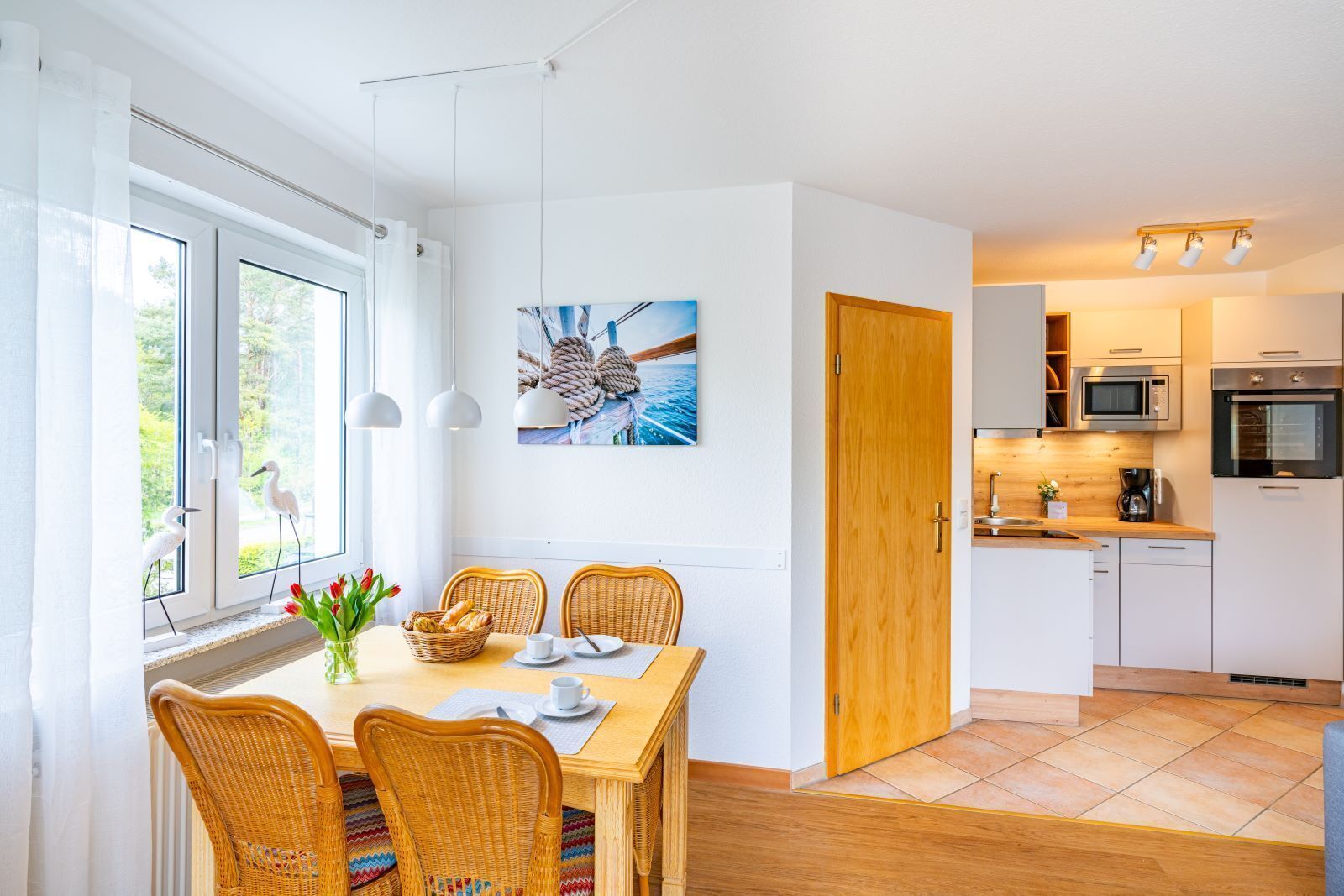 Een moderne keuken met witte kasten, een magnetron en een kookplaat. Er is een zithoek met rieten stoelen en een tafel, en een raam met gordijnen en een schilderij.