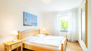 Une chambre à coucher bien rangée, avec un lit en bois, deux tables de chevet avec des lampes, une fenêtre ornée de rideaux légers et un tableau accroché au mur.