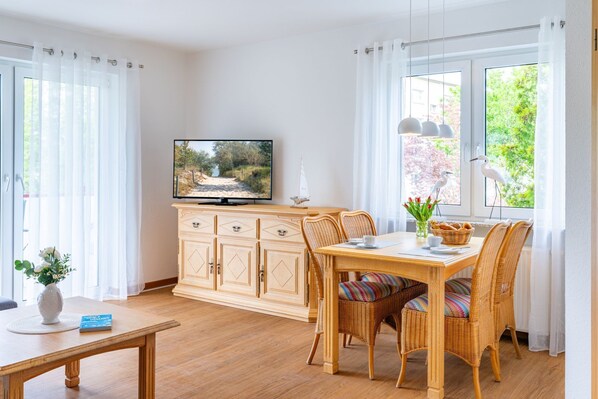 A dining area with a wooden table, wicker chairs, a television on a cabinet, and a view of greenery through the window.