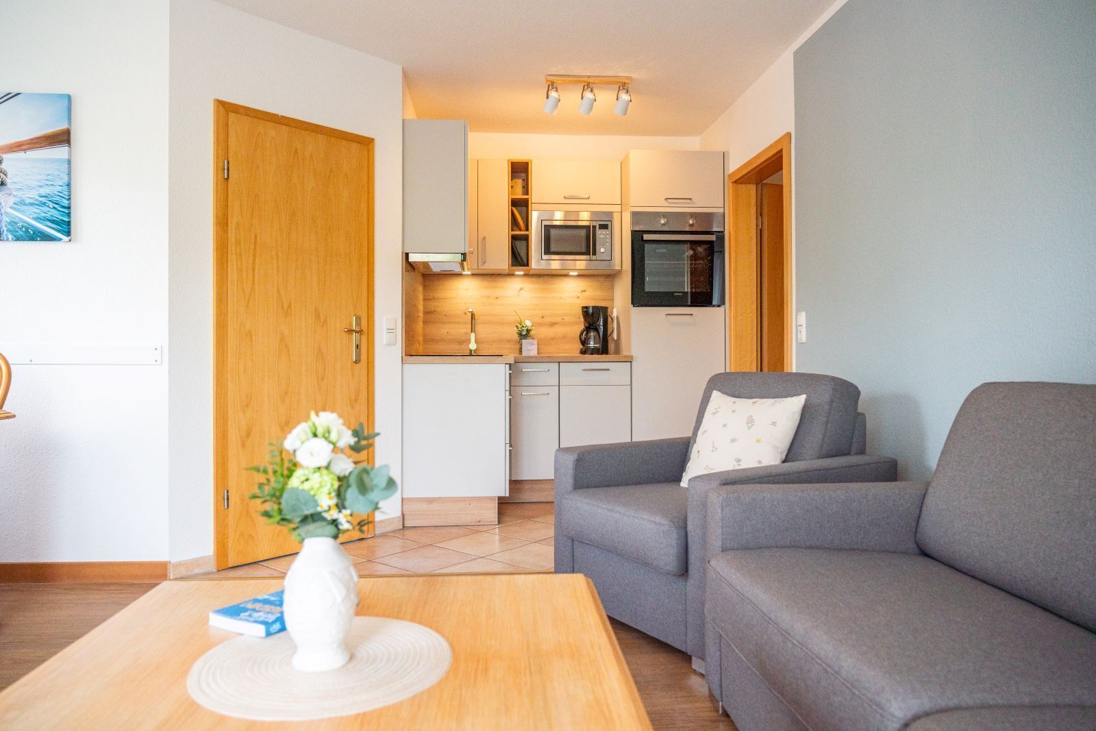 Een knus woongedeelte met een kitchenette, een grijze bank en een klein tafeltje met een vaas.