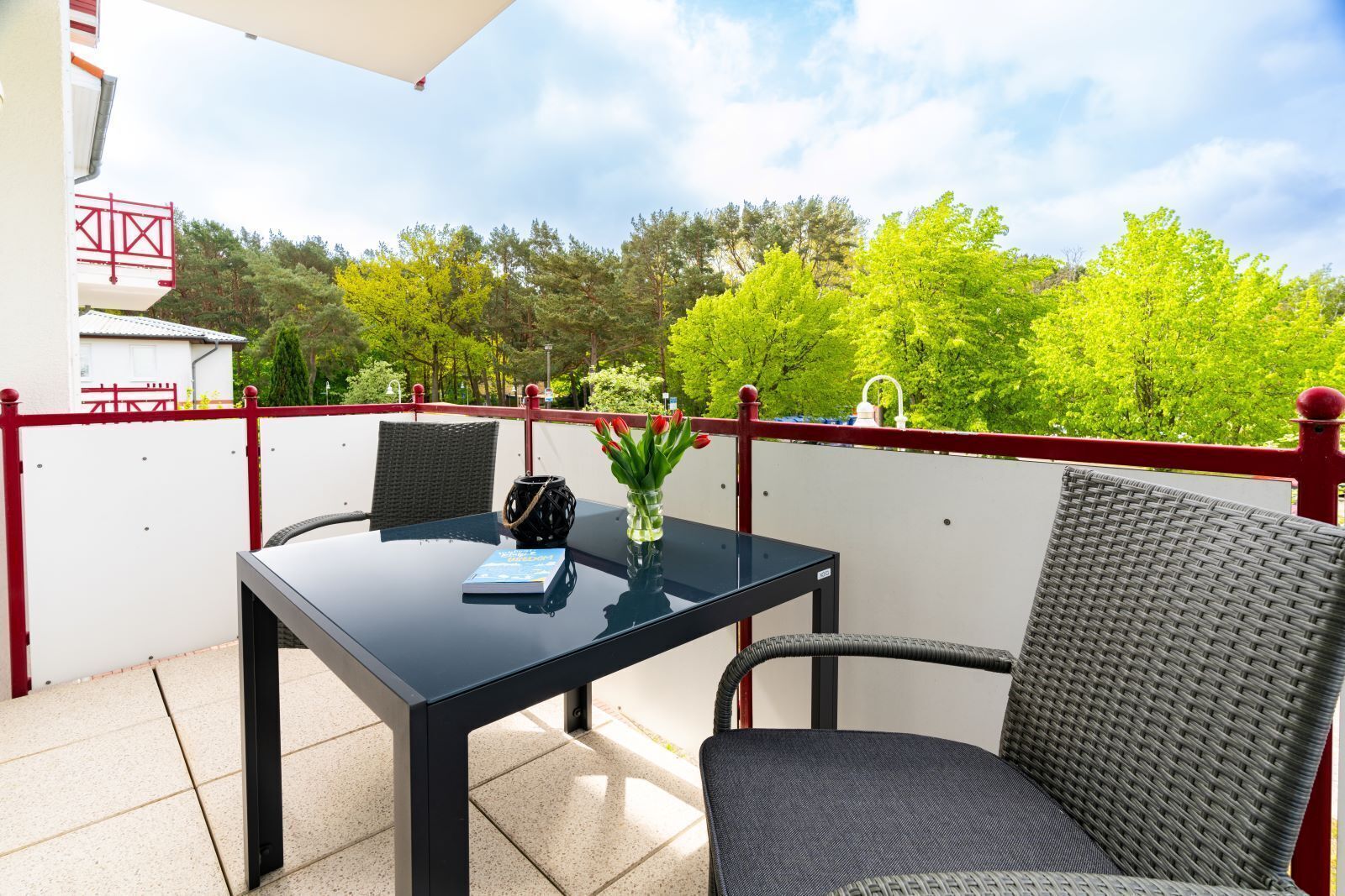 A balcony with a table and chairs, a vase with flowers, and a view of trees and a building in the background.