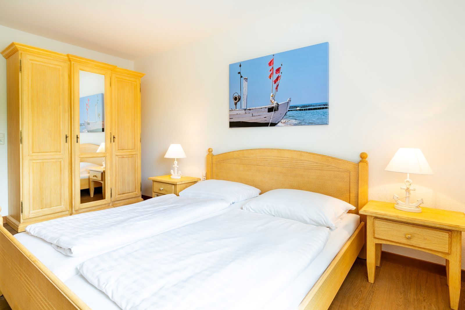 A bedroom with a bed, wooden wardrobe, bedside tables with lamps, and a painting of a boat on the wall.