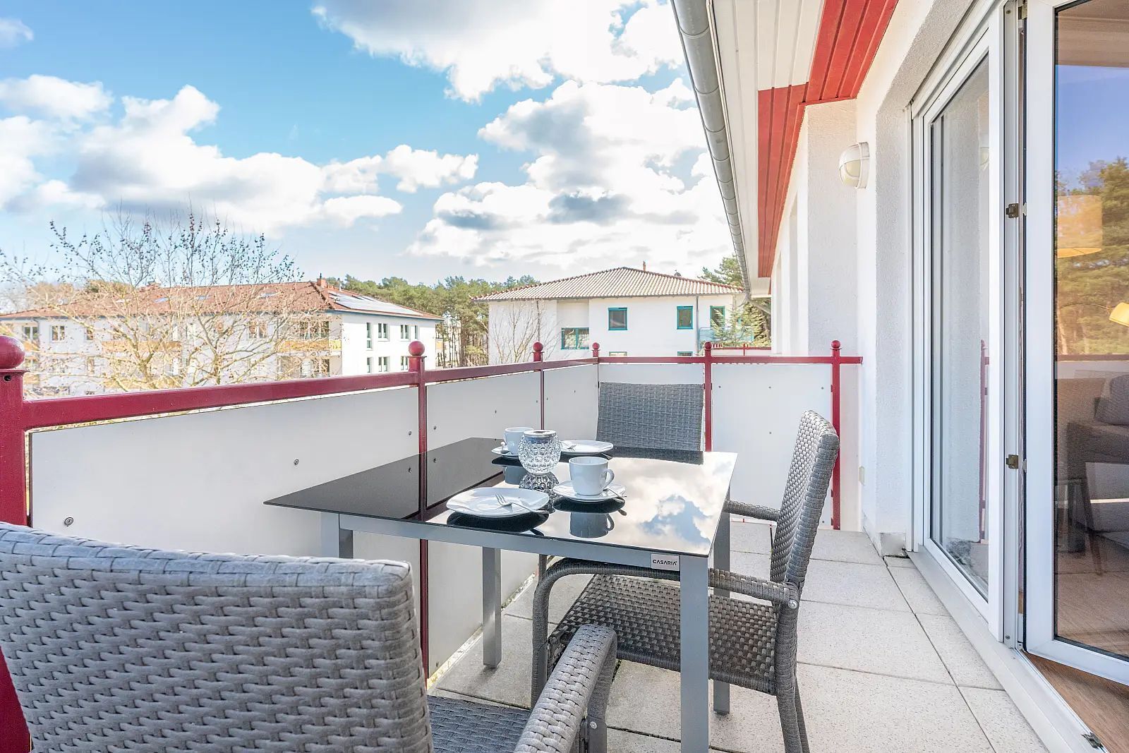 Un balcon avec une table dressée pour deux personnes, des chaises en osier et une porte vitrée donnant sur une pièce.