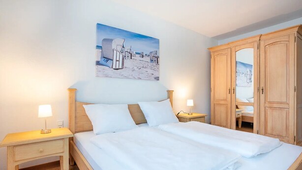 Un lit bien fait, avec du linge de lit blanc, deux tables de chevet avec des lampes, une armoire en bois et un tableau sur le thème de la plage accroché au mur.