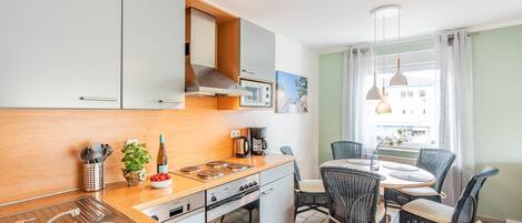 Cocina compacta con pileta, estufa y horno, comedor con mesa y sillas, y ventana con cortinas.
