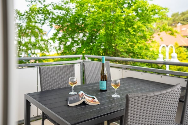 Et bord med en flaske og to glas, et magasin, samt fletstole på en udendørs balkon med gelænder og grønne planter i baggrunden.