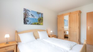 A neatly made bed with white linens, a wooden headboard, and two bedside tables with lamps. A large wooden wardrobe is to the right of the bed. - Meerliebe - Stern FeWo Strandstraße (Karlshagen)
